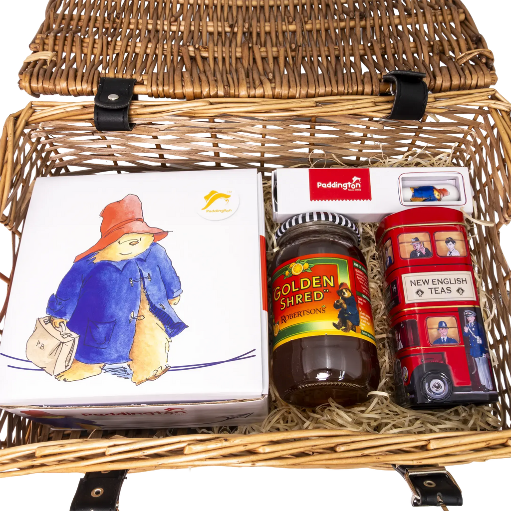 A wicker picnic basket filled with curated treats and toys on a white background. Visible items include a white Paddington box with a cartoon bear illustration, a jar of Golden Shred Pickled Ginger, a red double-decker bus toy, and a white tea box.