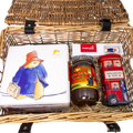 A wicker picnic basket filled with curated treats and toys on a white background. Visible items include a white Paddington box with a cartoon bear illustration, a jar of Golden Shred Pickled Ginger, a red double-decker bus toy, and a white tea box.