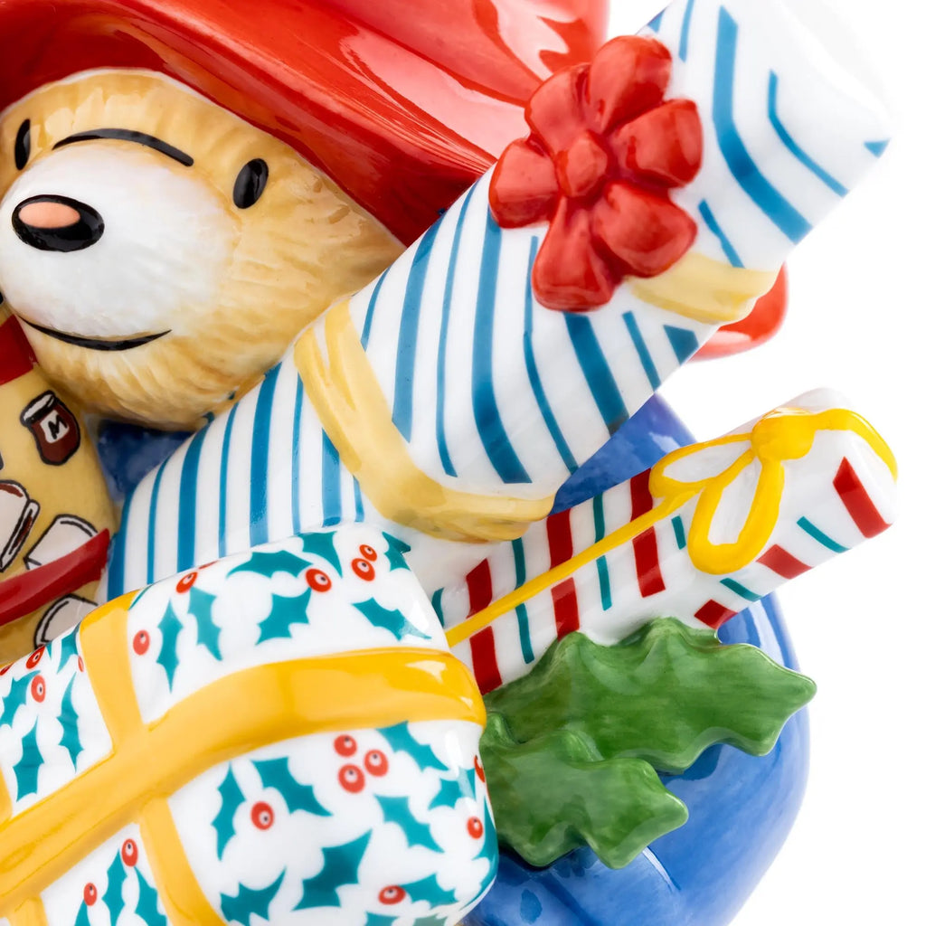 Close-up fine bone china figurine: bear face hugging striped gift bag with red bow.